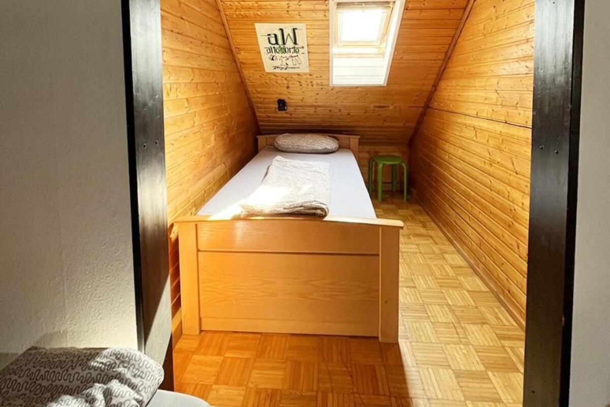 Un dormitorio abigado con techo inclinado de madera, una cama individual, un taburete pequeño y una ventana.