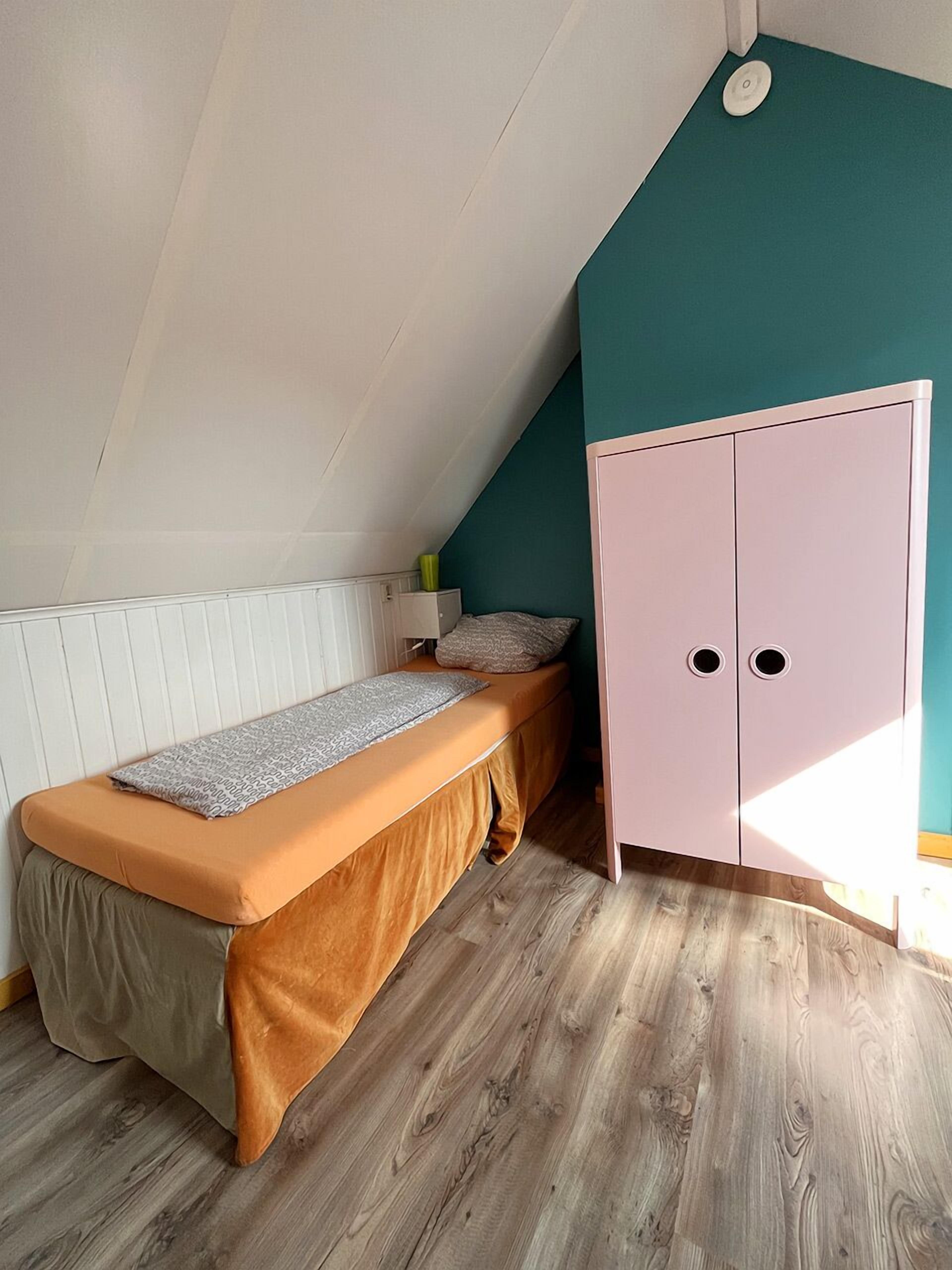 A bedroom with a sloped ceiling, a bed with a brown mattress and a grey blanket, a teal wall, and a pink wardrobe.