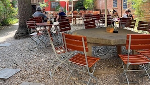 An outdoor seating area with tables and chairs under a green awning, surrounded by trees and a brick building. - Homeplanet Hostel (Leipzig)