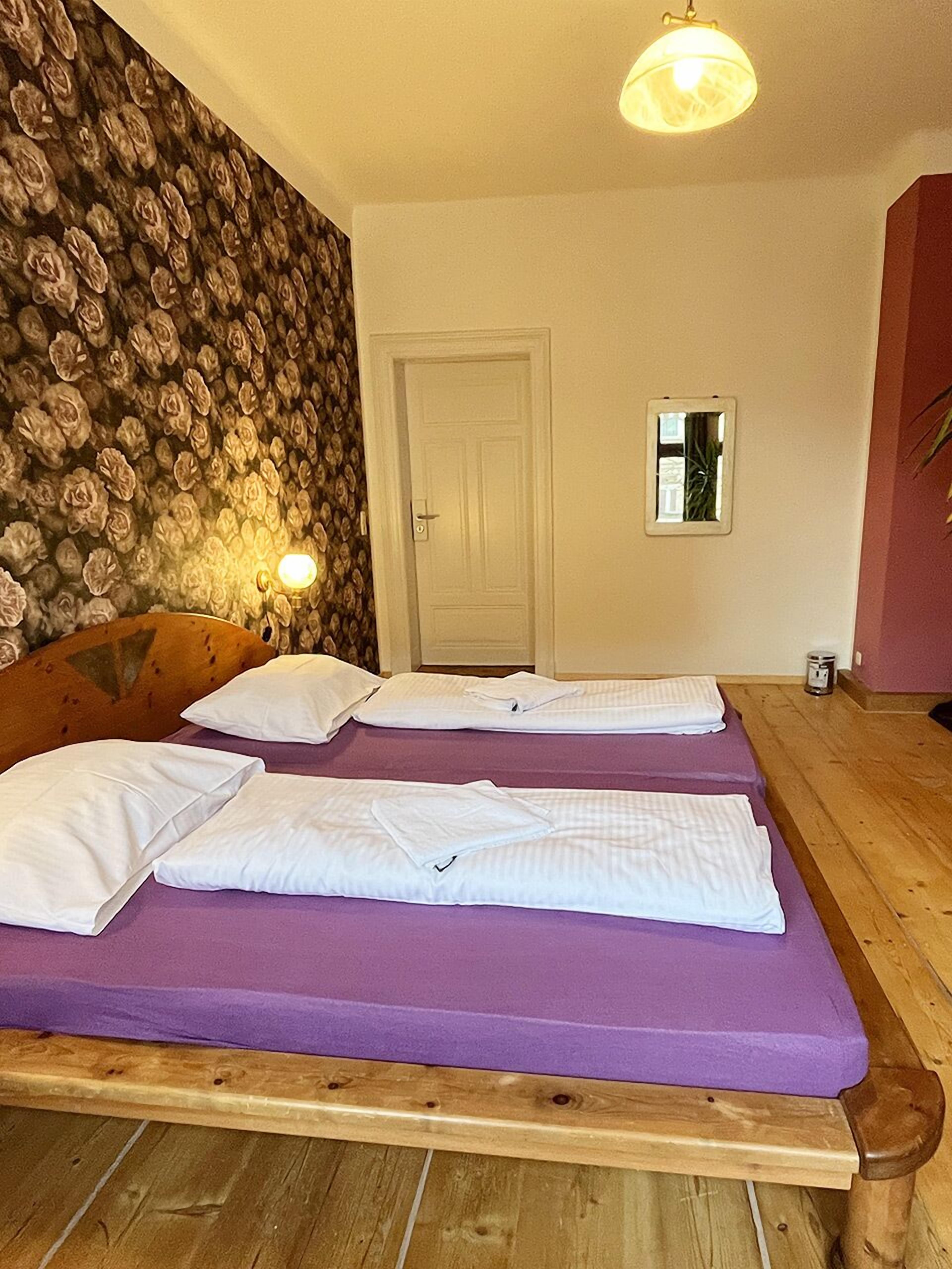 Ein Zimmer mit zwei Betten, einem hölzernen Kopfteil, einer blumengemusterten Wand, einer Tür und einem kleinen Fenster.