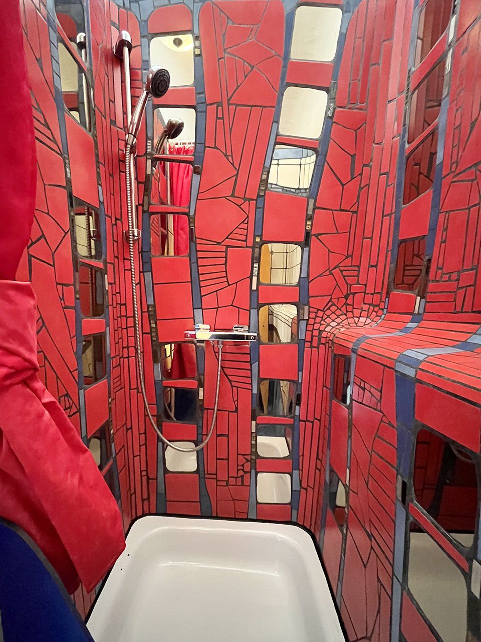 Ein Badezimmer mit Wandgestaltung aus roten und blauen Mosaiken, einer weißen Badewanne und einer Dusche mit Duschschlauch.
