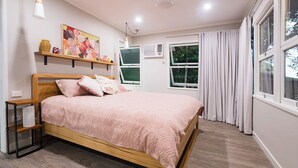 A bedroom with a wooden bed, a bedside table, a wall-mounted shelf with decorative items, a window with curtains, and an air conditioning unit. - The Point. A rainforest and ocean view retreat (Flying Fish Point)