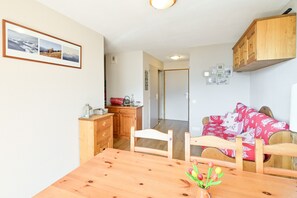 A dining area with a wooden table, chairs, a vase with flowers, a kitchen area with wooden cabinets, and a hallway leading to a bedroom with a bed and red bedding. - Apartment for 6 in Saint François Longchamp (1650 m) (Saint-François-Longchamp)