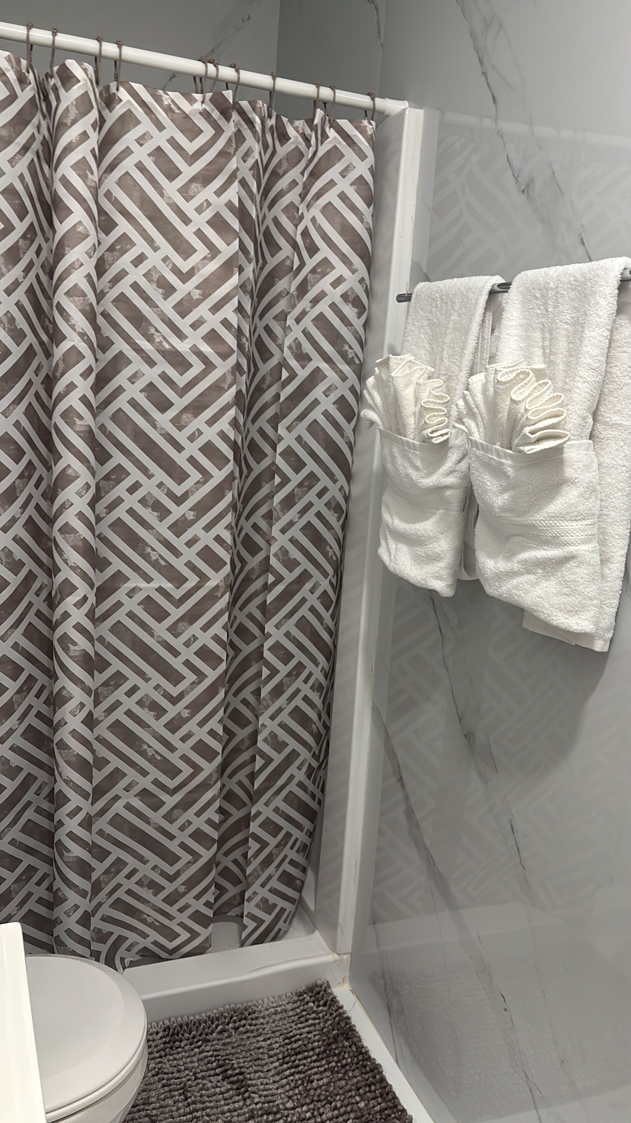 Une salle de bain avec un rideau de douche orné d’un motif géométrique, un porte-serviettes blanc avec des serviettes et un mur en marbre.
