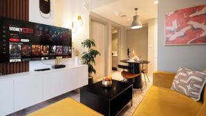 A modern living room with a large flat-screen TV displaying a streaming service interface, a black coffee table with fruit, a yellow sofa, and a dining area with a small table and chairs. - Lake-View 1BR Retreat in Business Bay  (Dubai)