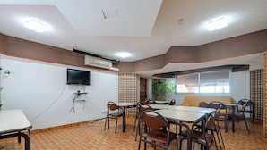Un comedor con suelo de baldosas, mesas, sillas, un sofá, un televisor montado en la pared y un equipo de aire acondicionado.