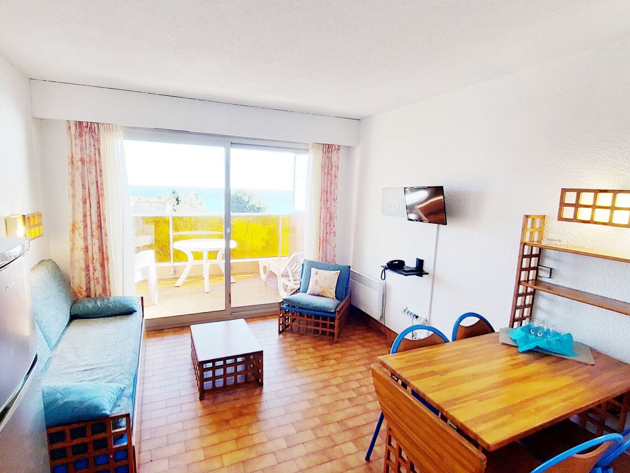 A room with a wooden floor, a sofa, a table, chairs, a TV, and a balcony with a table and chairs.