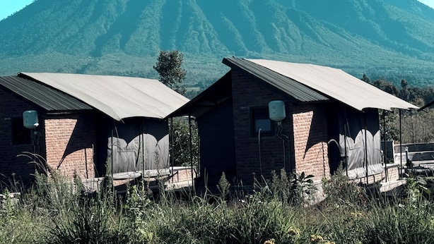 Two brick buildings with metal roofs, likely cabins or small lodges, set against a backdrop of a large mountain. - Volcanoes Over View Safaris & Camp  (Kinigi)