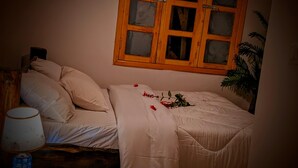 A bedroom with a bed, a nightstand, a lamp, and a window with a wooden frame. - Volcanoes Over View Safaris & Camp  (Kinigi)