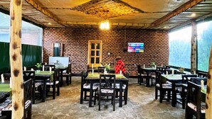 A restaurant with brick walls, wooden tables and chairs, a TV screen, and a stone floor. - Volcanoes Over View Safaris & Camp  (Kinigi)