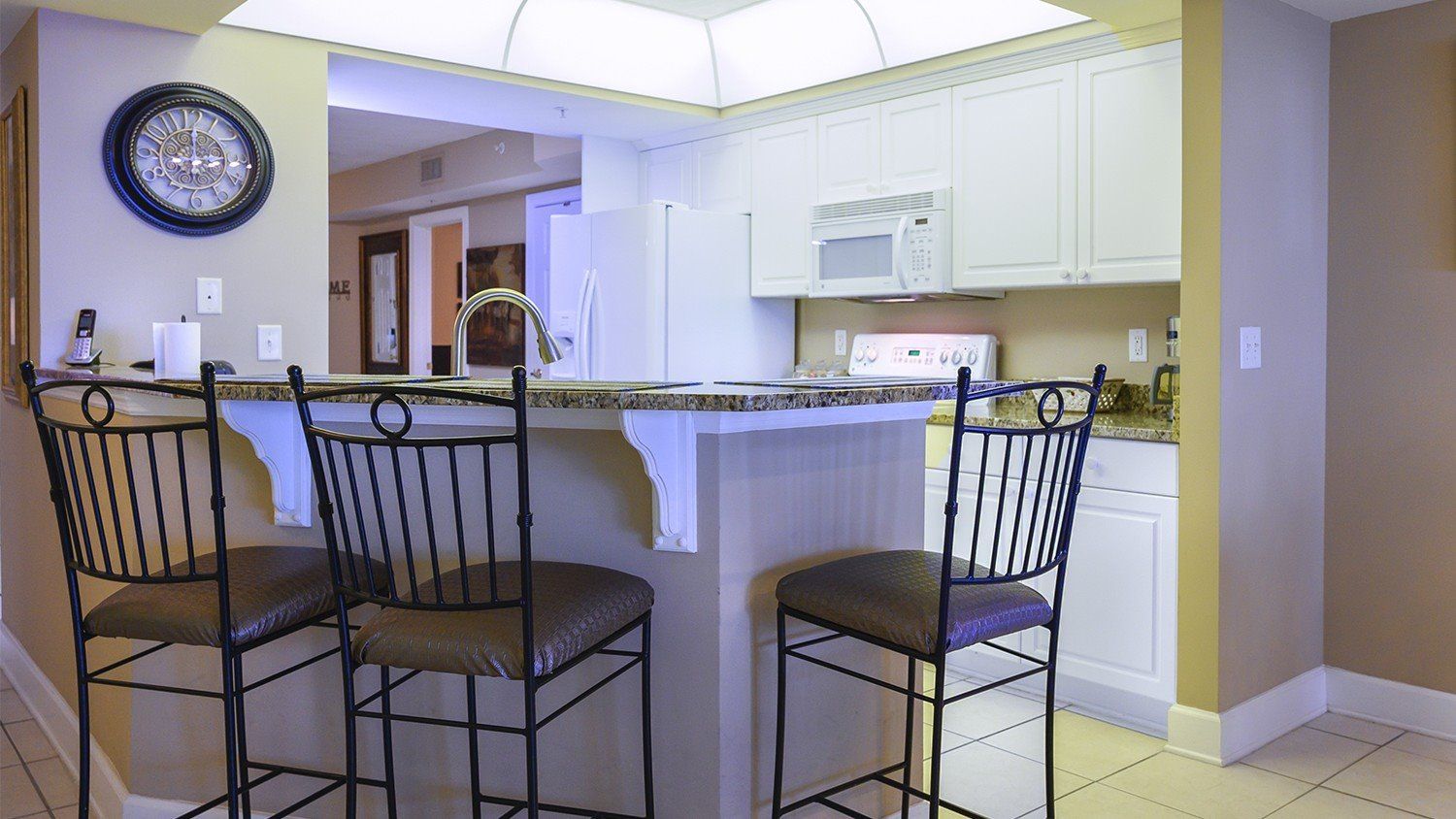 A kitchen with a bar area, white cabinets, a microwave, and a clock on the wall.