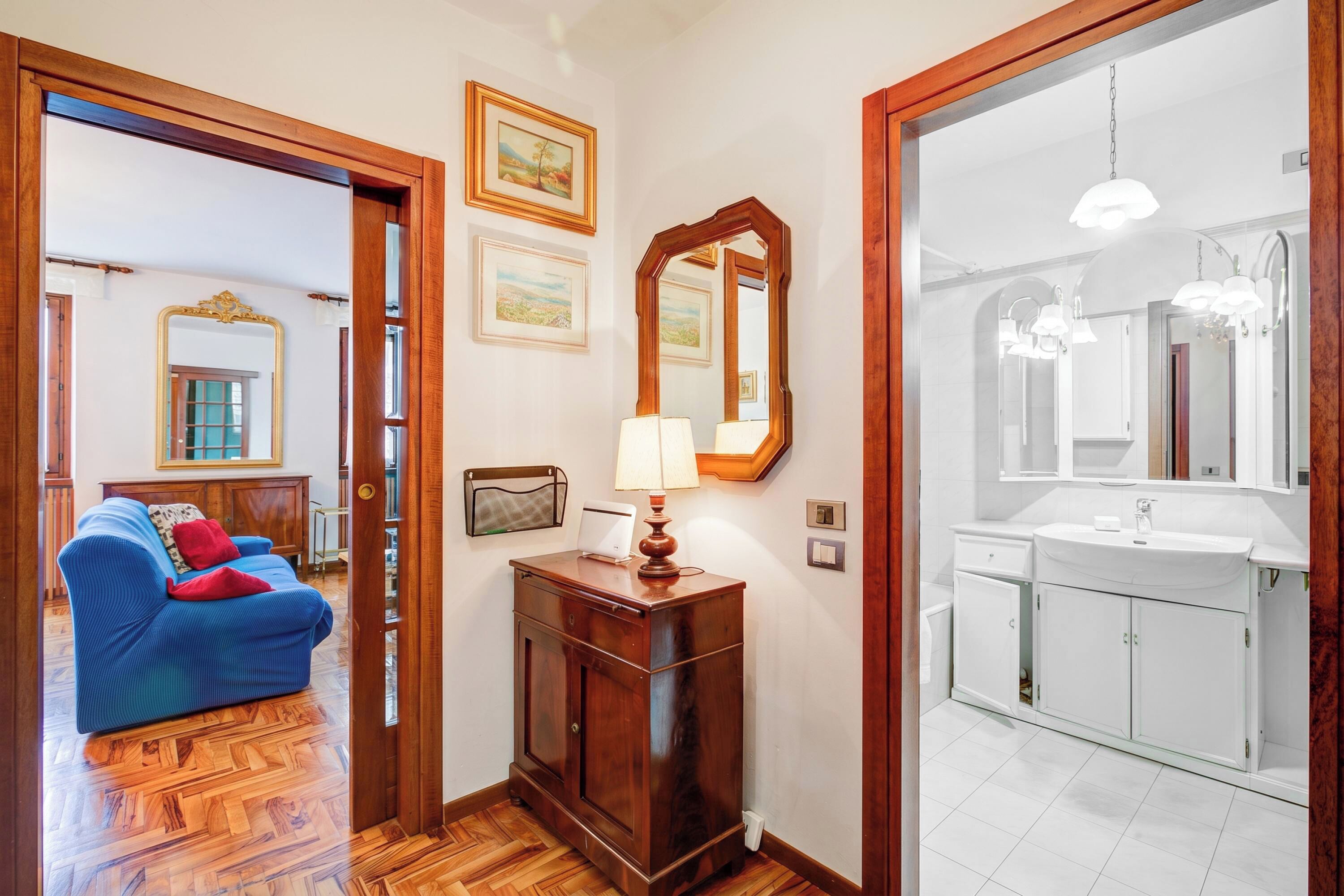 Une chambre d’hôtel avec un plancher en bois, un canapé bleu, une armoire en bois avec une lampe, et une salle de bain avec un lavabo et un miroir.