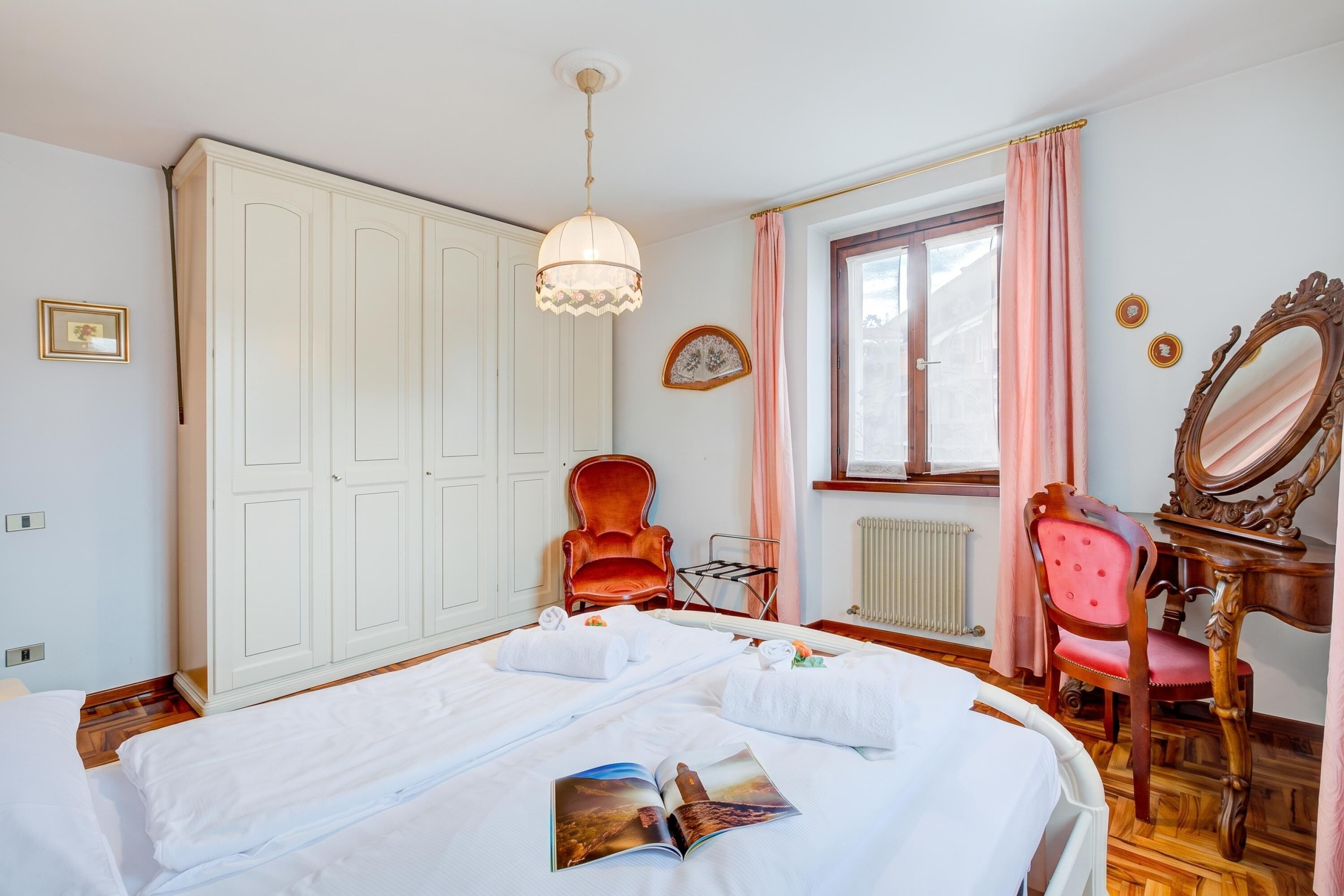 Une chambre d’hôtel avec un lit, une chaise, une coiffeuse avec un miroir, une fenêtre avec des rideaux et un radiateur.