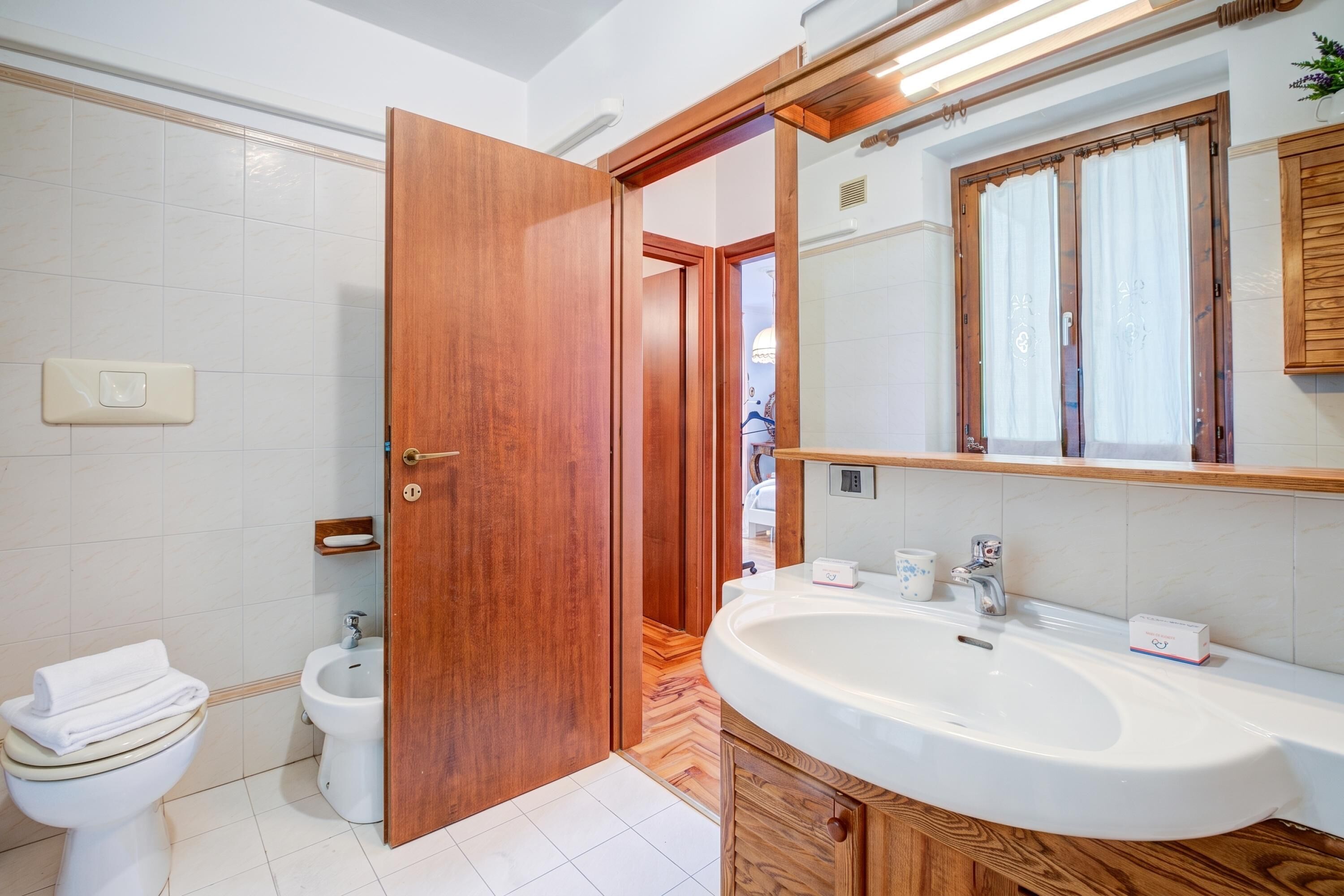 Un baño con puerta de madera, azulejos blancos, lavamanos blanco y un inodoro.