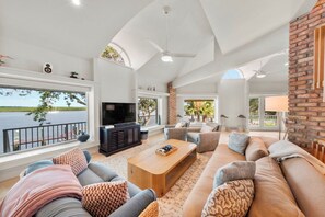 A spacious living room with a large TV, comfortable seating, and a view of the water.