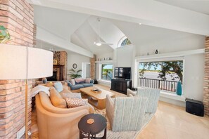 A spacious living room with a brick accent wall, a fireplace, a large TV, and a balcony with a view. - Ponce Inlet Waterfront Escape - 4 Bedroom 3 Bath (Ponce Inlet)