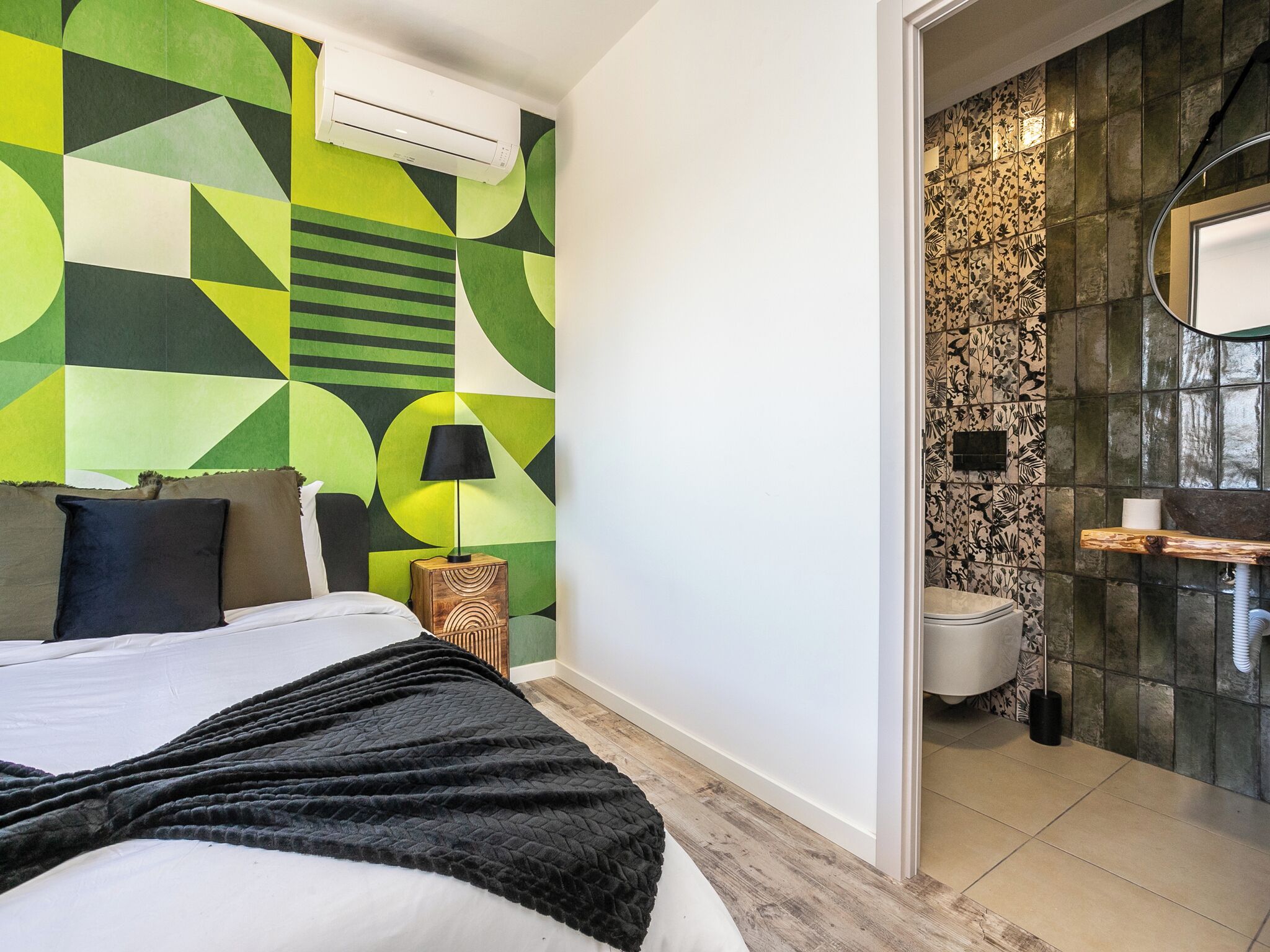 Ein modernes Hotelzimmer mit Wand im geometrischen Muster, einem Bett mit schwarzer Decke, einer Nachttischlampe und einem kleinen Nachttisch aus Holz.
