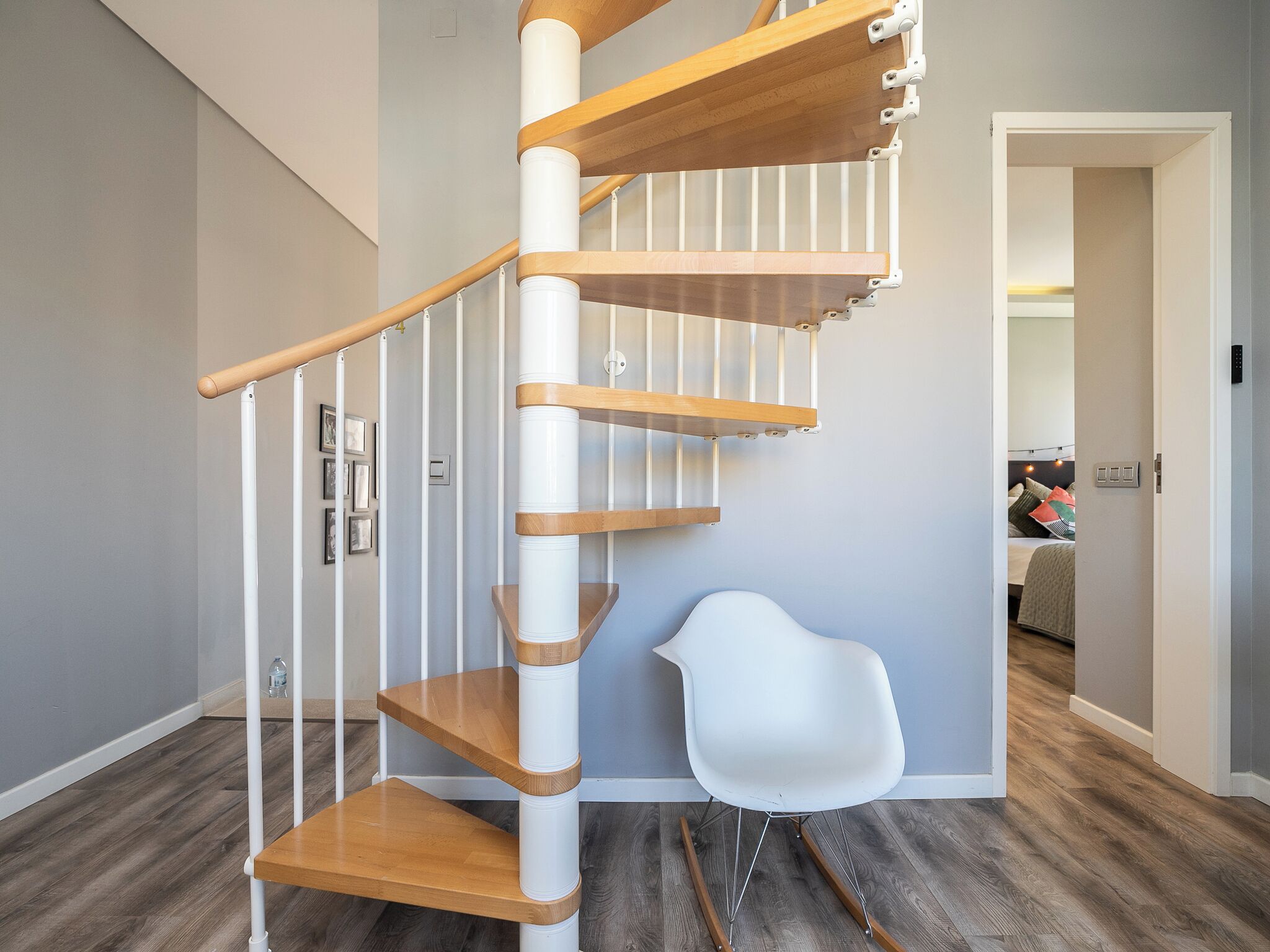 A modern interior with a wooden spiral staircase, a white chair, and a visible bedroom with a bed and pillows.