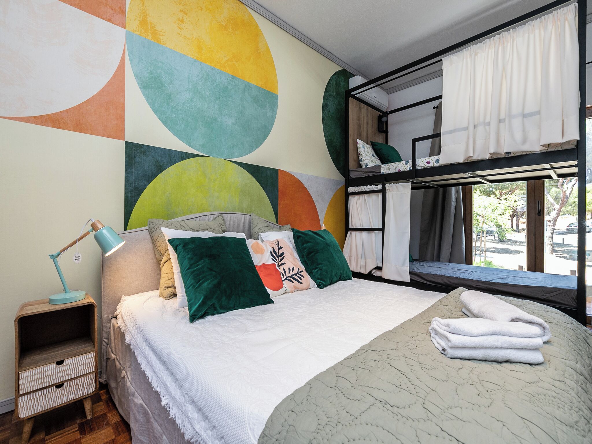 A bedroom with a bunk bed, a bedside table with a lamp, and a large abstract wall mural.