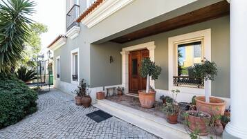 Ein Kopfsteinpflasterhof mit einem Gebäude, das eine Holztür, große Pflanzgefäße und einen Balkon mit Geländer zeigt.