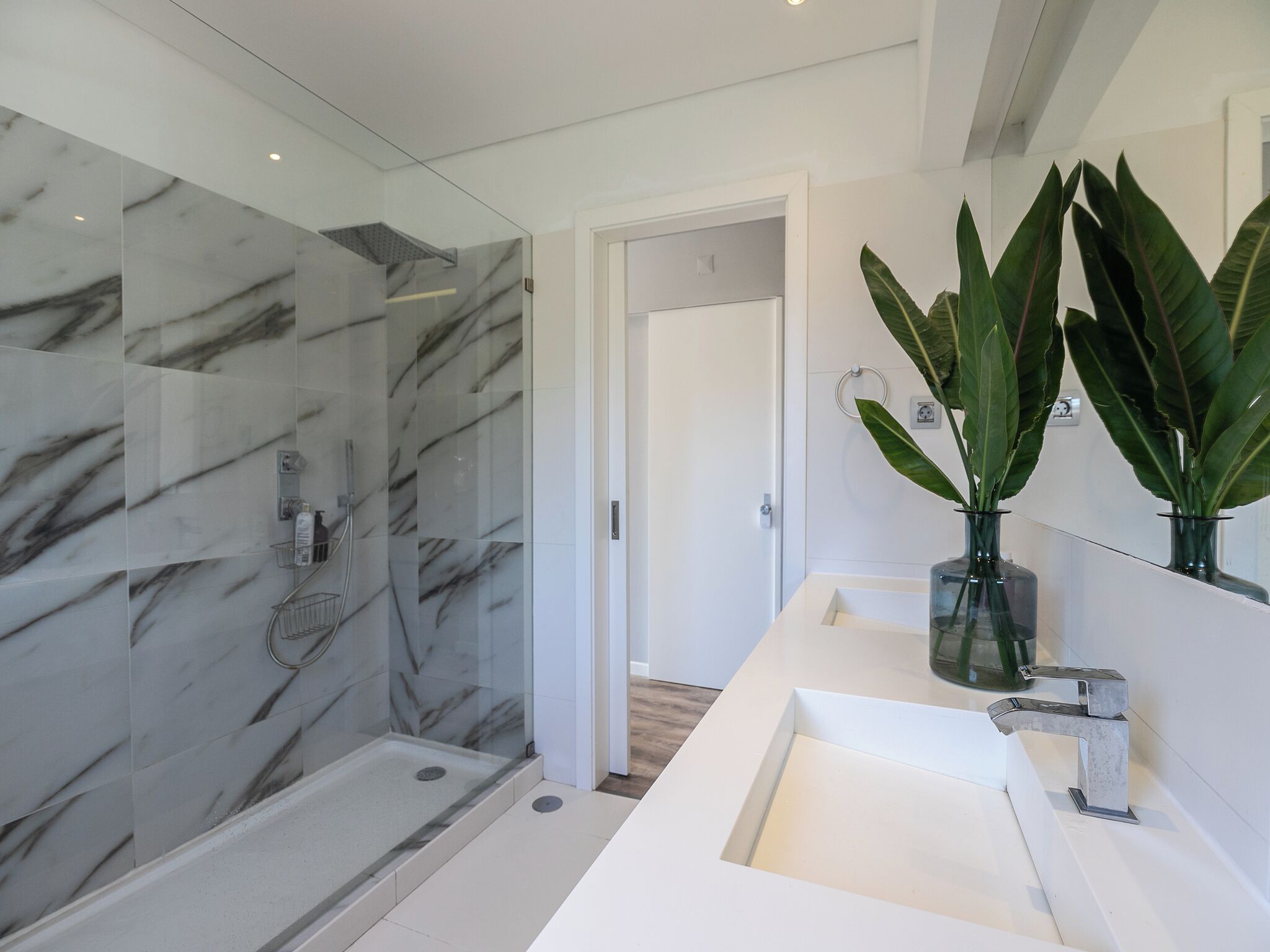 A modern bathroom with a glass-enclosed shower, a white sink, and two large green plants.
