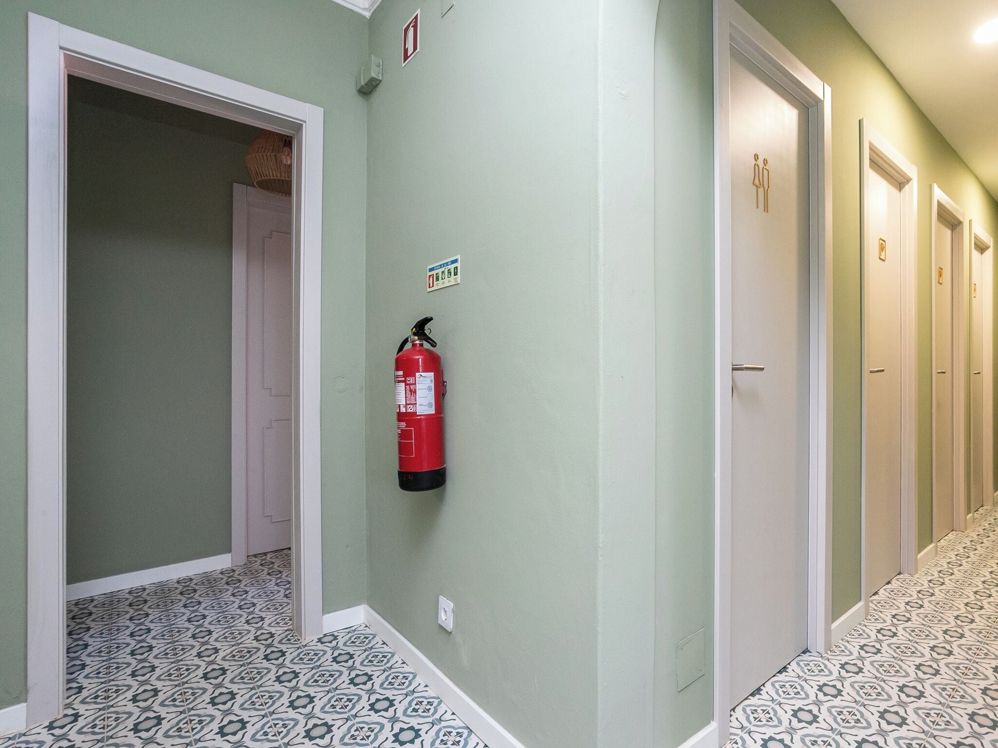 A hallway with patterned floor tiles, a fire extinguisher mounted on the wall, and doors leading to individual rooms.