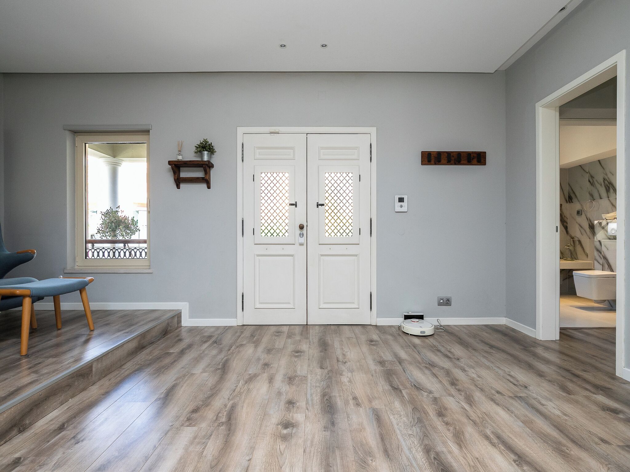 Amplia sala de estar con suelo de madera, un doble acceso con panel de vidrio decorativo, un pequeño estante con una planta y vistas a un balcón a través de una ventana.