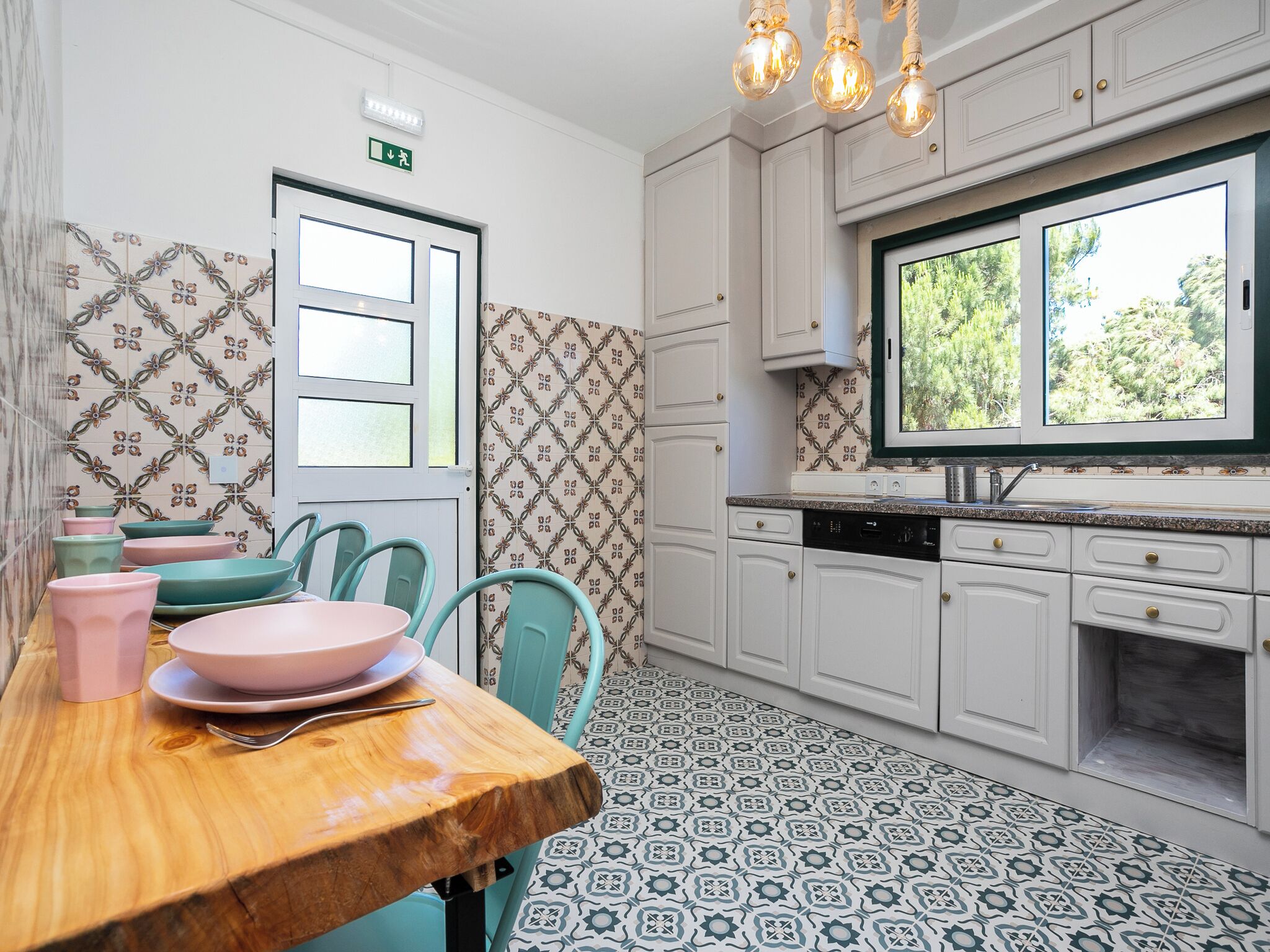 A kitchen with a wooden table, teal chairs, white cabinets, patterned floor tiles, and a window with a view of trees.