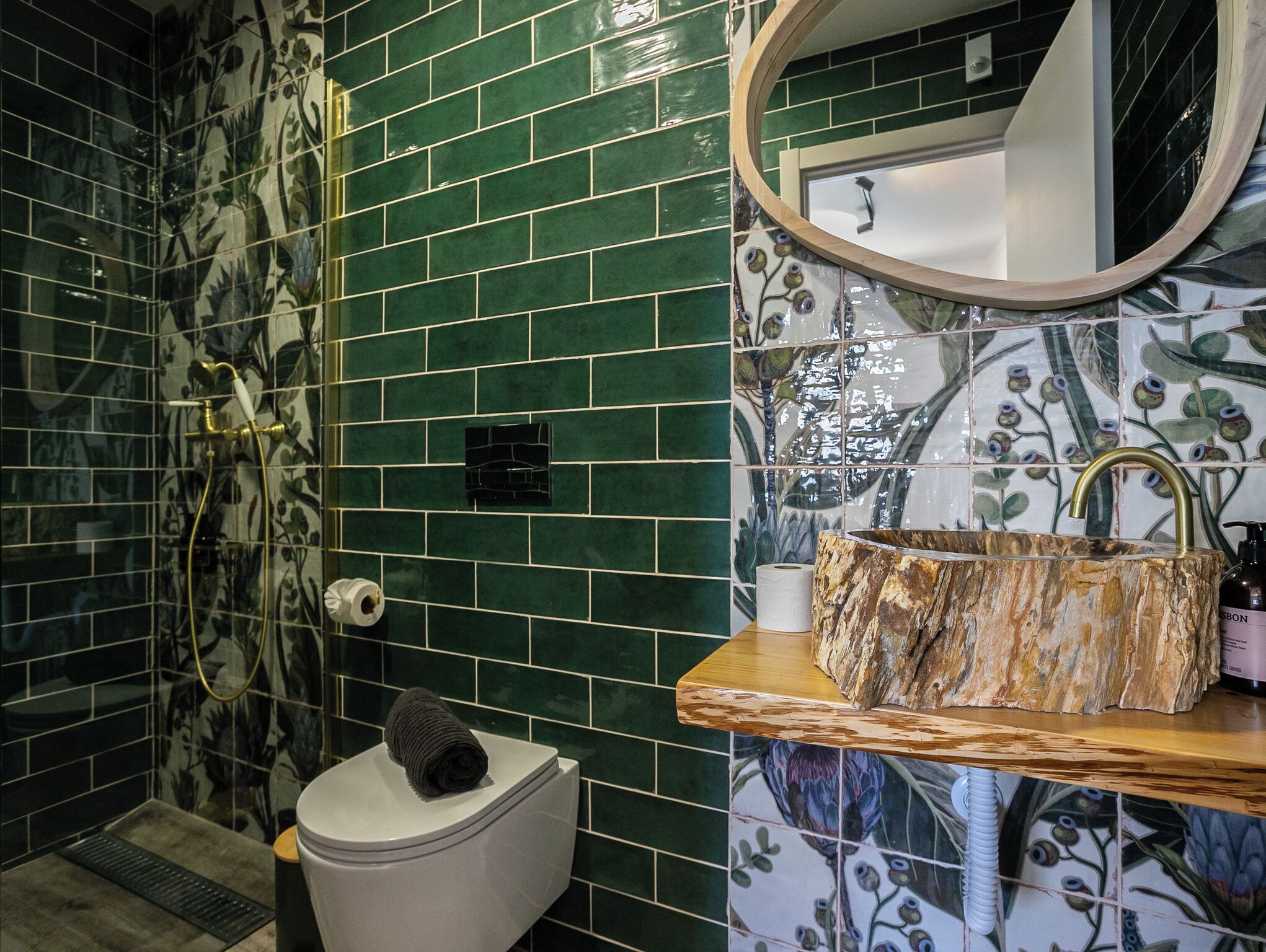 Baño con azulejos florales verdes, un inodoro, una ducha con grifo dorado y un lavabo con encimera de madera.