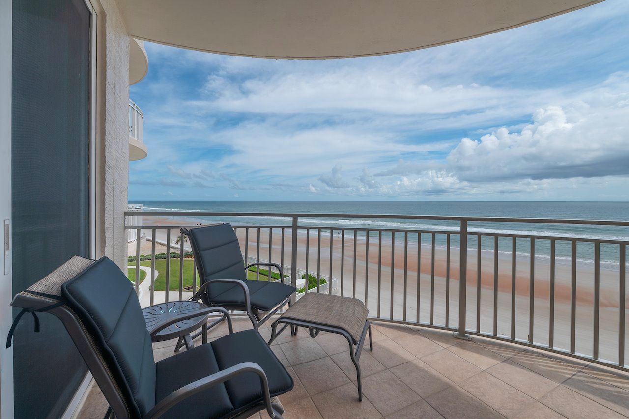 A balcony with two chairs and a small table, overlooking a beach with a clear sky.