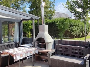 An outdoor patio with a stone pizza oven, a table with a tablecloth, and a wooden bench.