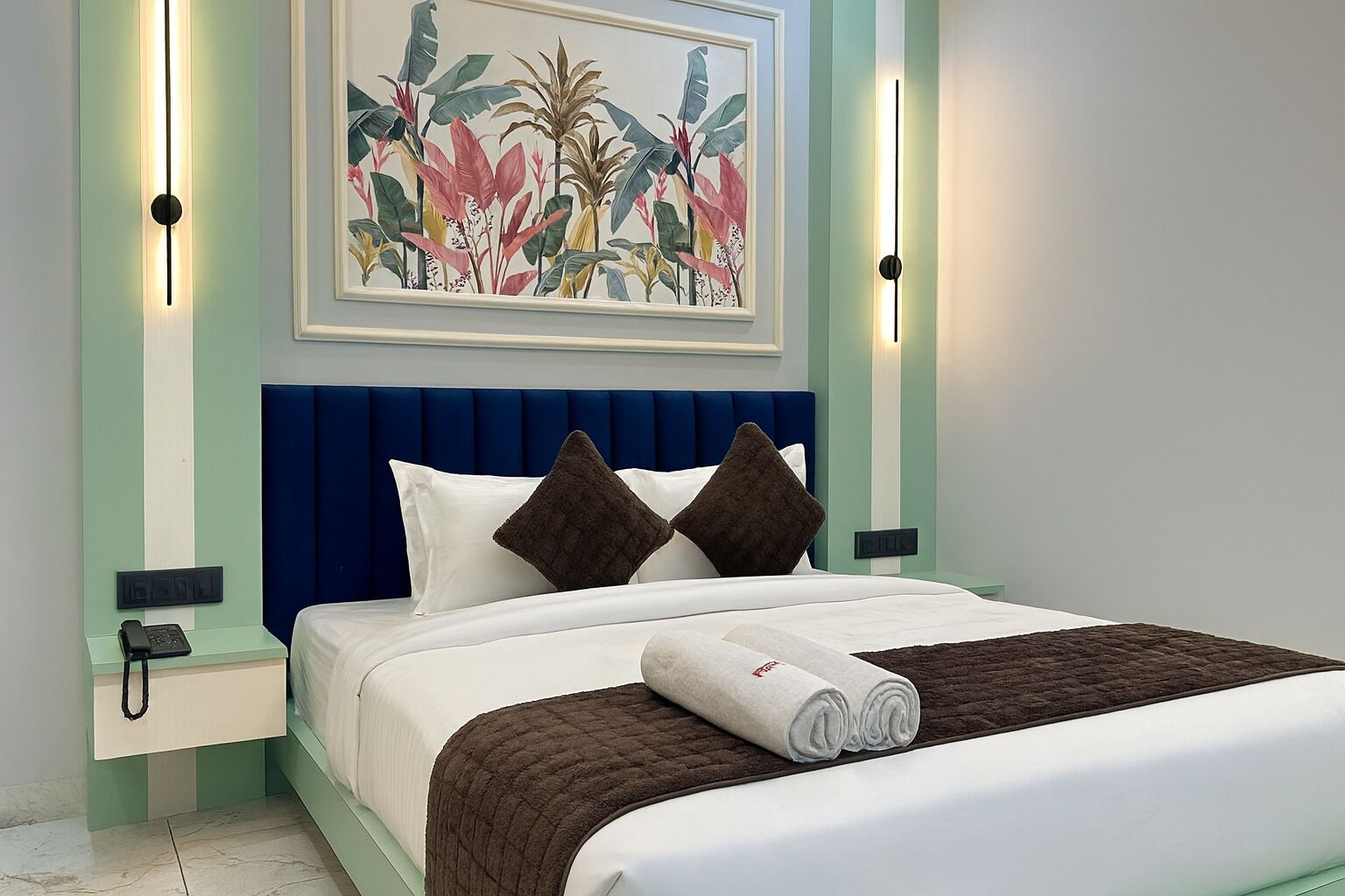 A neatly made bed with a blue headboard, white bedding, and brown pillows. There is a framed botanical artwork above the bed, and bedside lamps on either side.