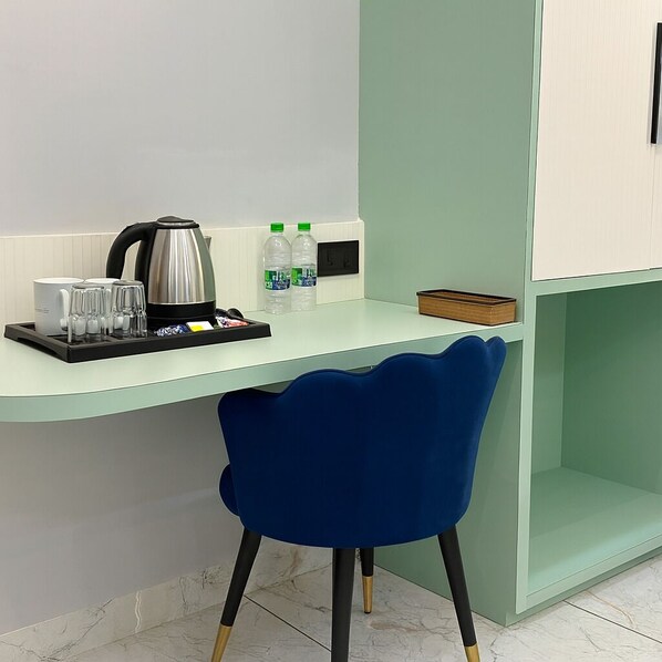 A modern hotel room with a curved desk, a blue chair, a kettle, glasses, and bottled water.