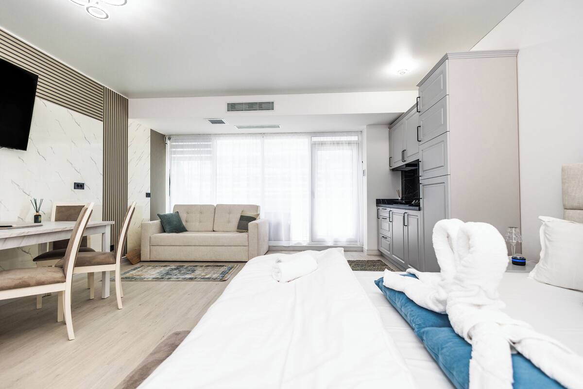 A modern hotel room with a bed, a sofa, a dining table with chairs, a TV mounted on the wall, and a kitchen area.