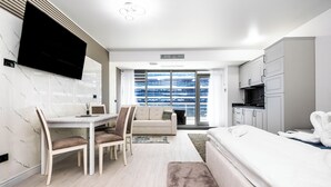 Ett modernt hotellrum med en platt-TV som är monterad på väggen, en matplats med bord och stolar, en soffa och en säng med ett vitt täcke.