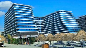 Ett modernt hotell med en böjd fasad, flera balkonger och ett läge vid stranden, med parasoller av halm.