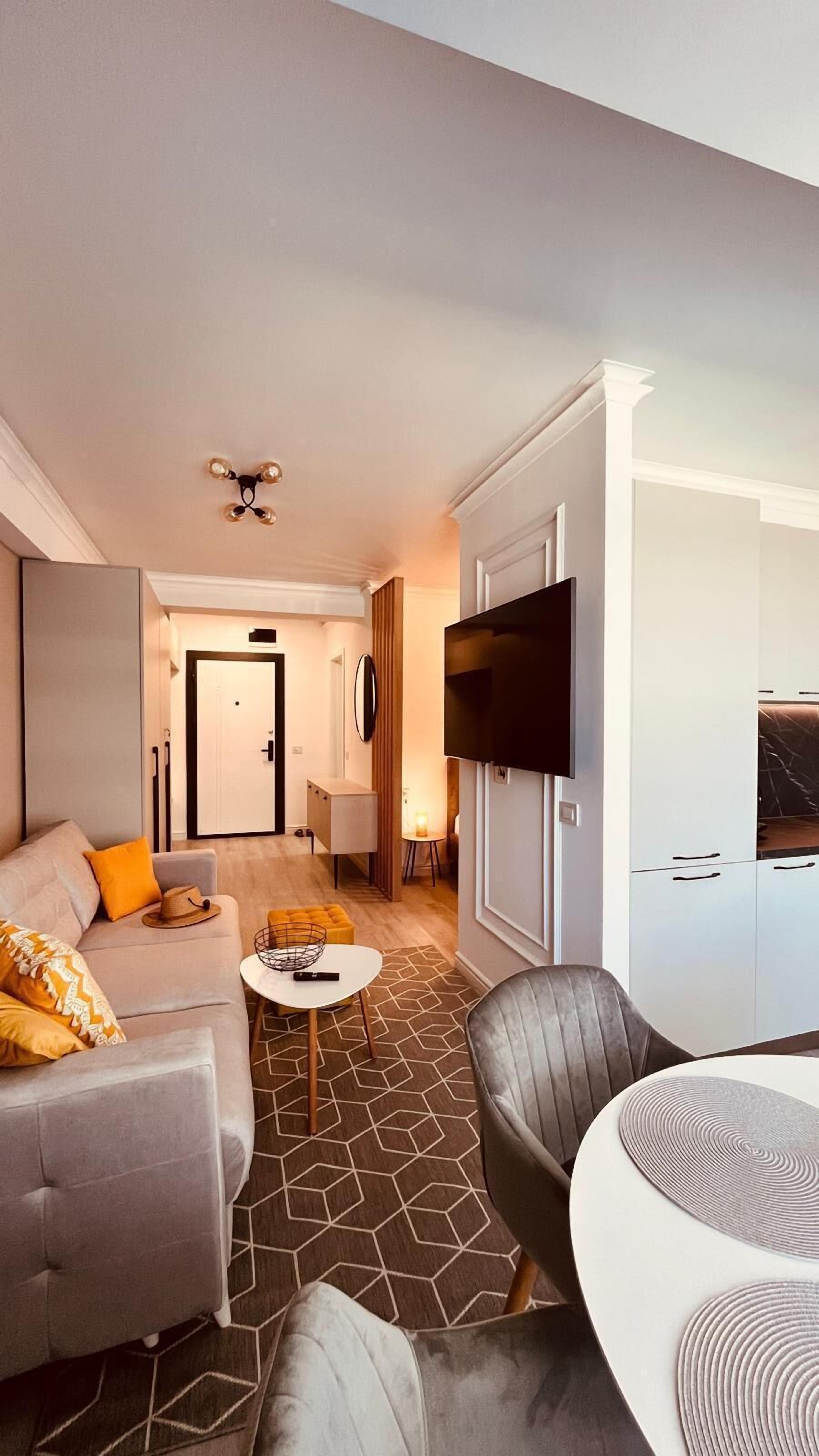 Ein modernes Hotelzimmer mit grauem Sofa, einem kleinen runden Tisch, einem grauen Sessel, einem an der Wand befestigten Fernseher und einer Tür zum Flur.