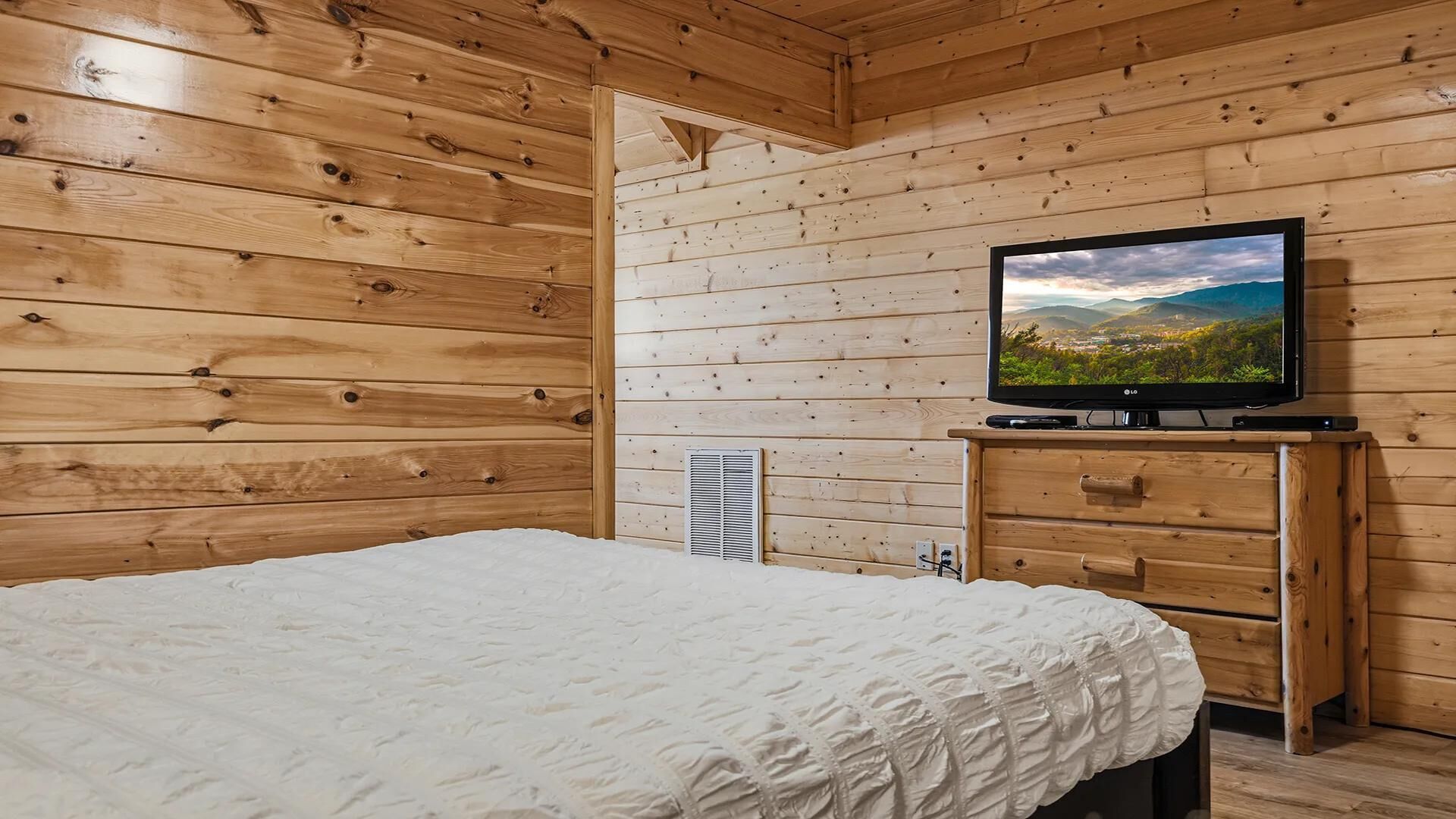 A wooden cabin room with a bed, a TV on a wooden stand, and a vent on the wall.