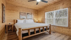 Ein Schlafzimmer einer Holzhütte mit Bett, Nachttisch, Deckenventilator und großem Fenster mit Jalousien.