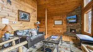 Interior acogedor de una cabaña con chimenea de piedra, paredes de madera, un sofá, una mesa de centro y un televisor colocado sobre la chimenea.
