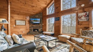 Interior acogedor de una cabaña con paredes de madera, una chimenea de piedra, un televisor y grandes ventanas con vistas al bosque circundante.