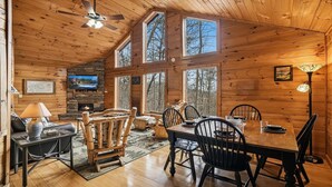 Interior acogedor de una cabaña con chimenea de piedra, una mesa de comedor de madera y grandes ventanales con vistas al bosque circundante.