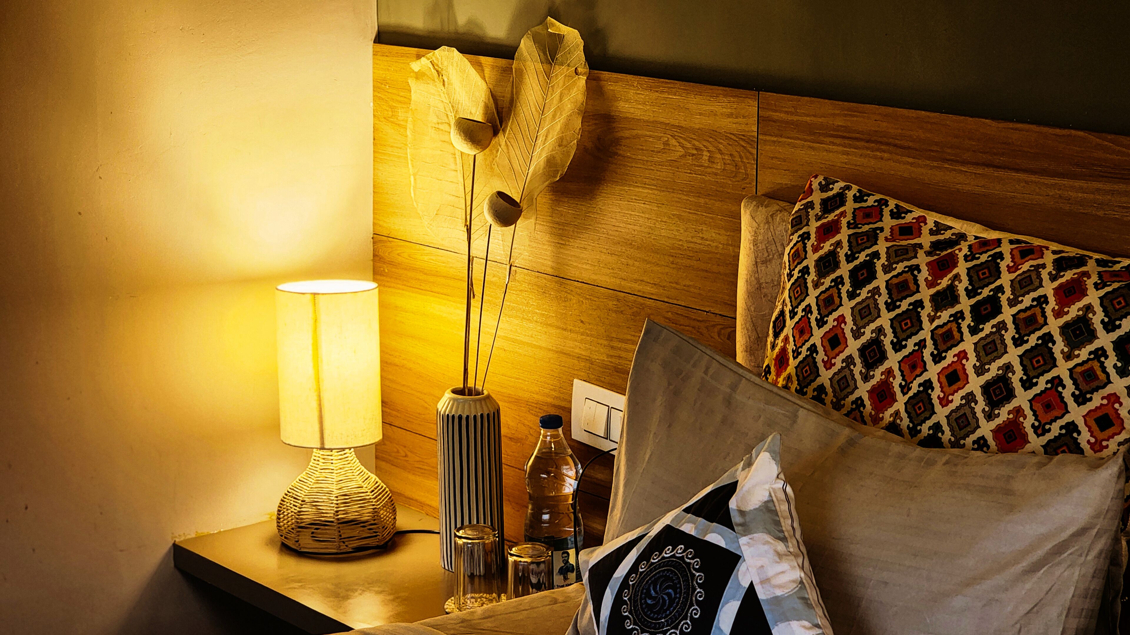 Habitación de hotel ordenada con cabecera de madera, lámpara de noche, una botella y un cojín estampado.
