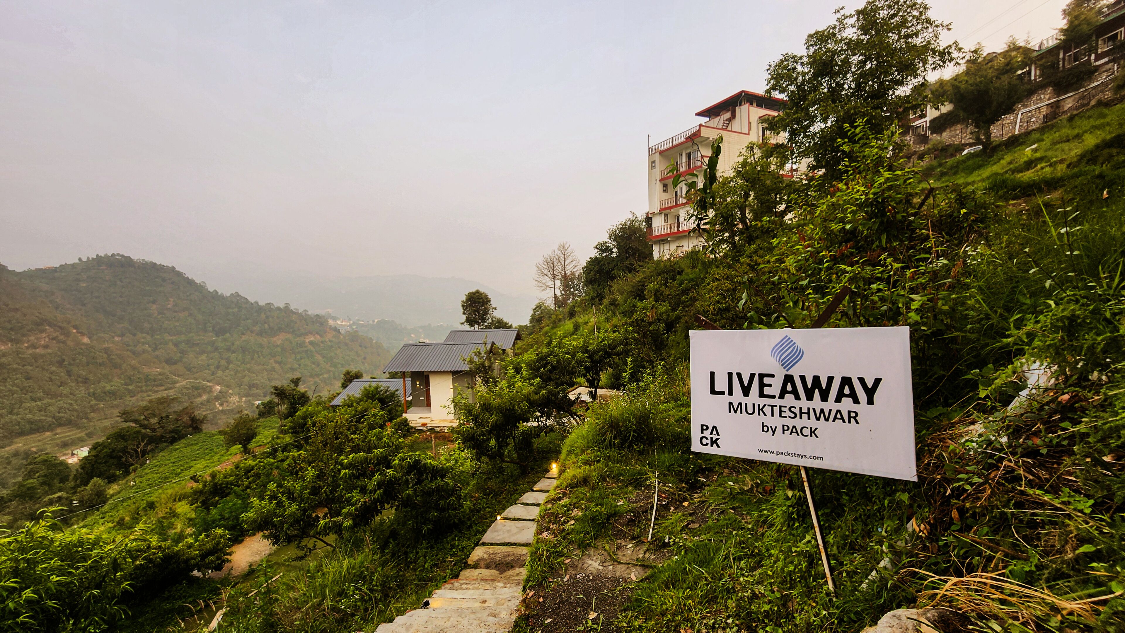 Een bordje voor LiveAway Mukteshwar by Pack, dat een accommodatie in een heuvelachtig gebied met weelderige begroeiing en uitzicht op verre heuvels aangeeft.