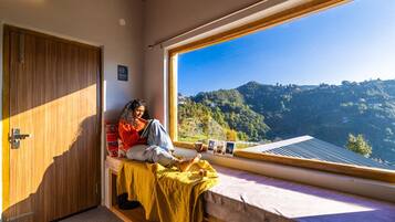 A room with a bed, a window with a view of a hilly landscape, a wooden door, and a small table with a cup and pictures.