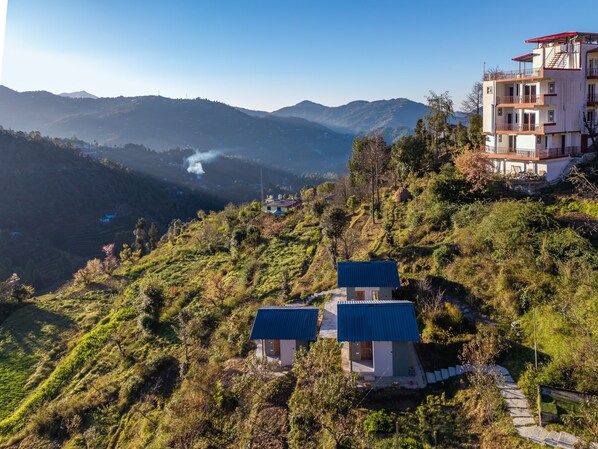 Una ladera con dos edificios pequeños con techos azules, un edificio más grande con balcones, y una vista de montañas distantes.
