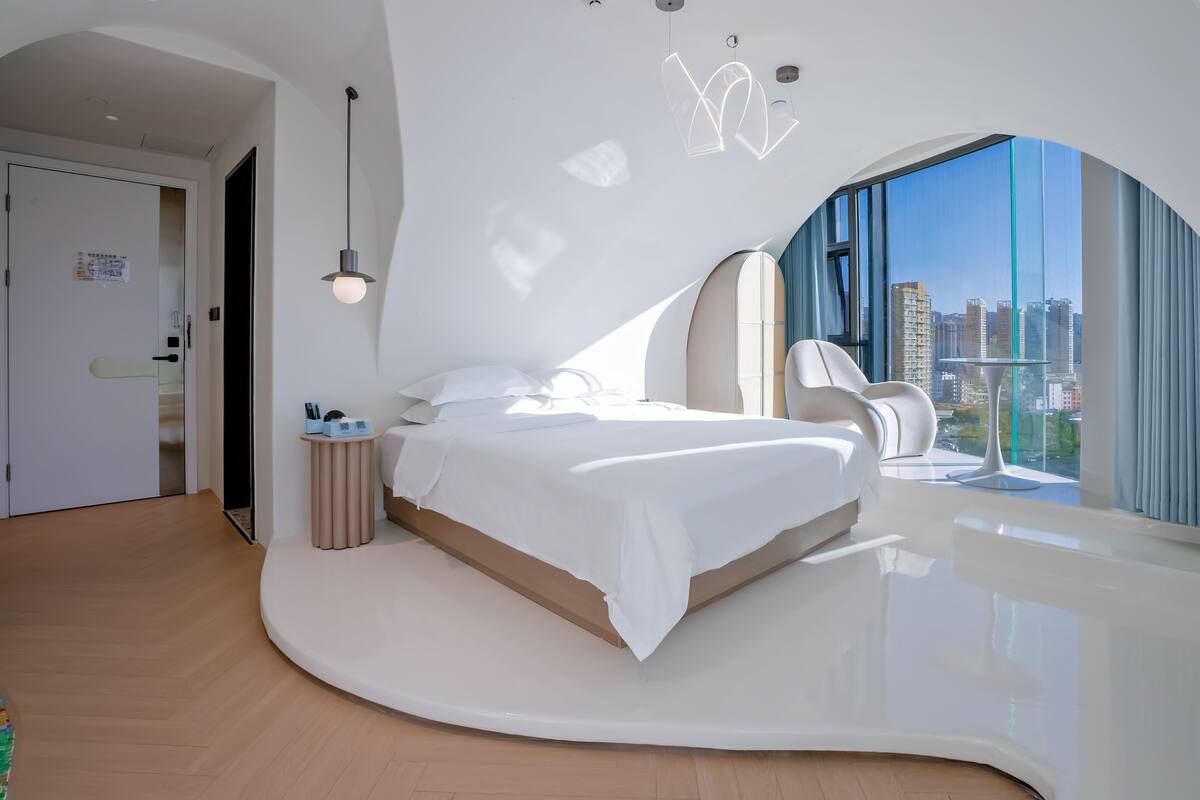 Un dormitorio moderno con techo curvo, una cama, una mesita de noche y vistas a la ciudad a través de grandes ventanales.