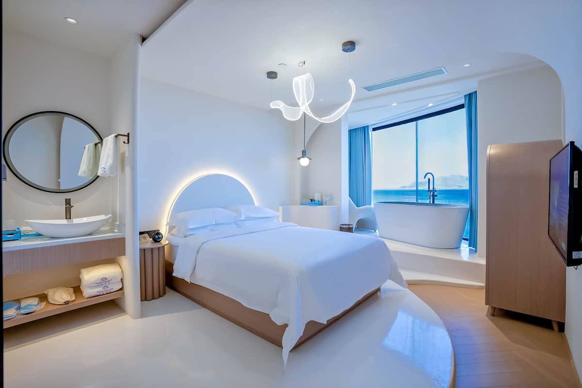 Una habitación de hotel moderna con una cama grande, un espejo redondo, un baño con bañera y vista al mar.