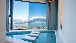 Une piscine intérieure moderne, avec des carreaux bleu clair, offrant une vue sur le paysage urbain et les montagnes à travers de grandes fenêtres, et un simple banc blanc le long du bord de la piscine.