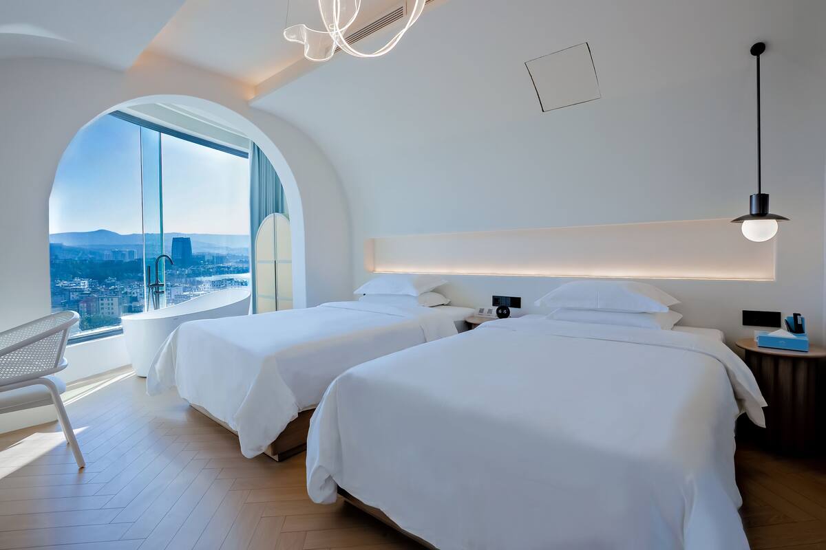 Una habitación de hotel moderna con dos camas, un amplio ventanal de arco, vistas a la ciudad y un diseño minimalista.